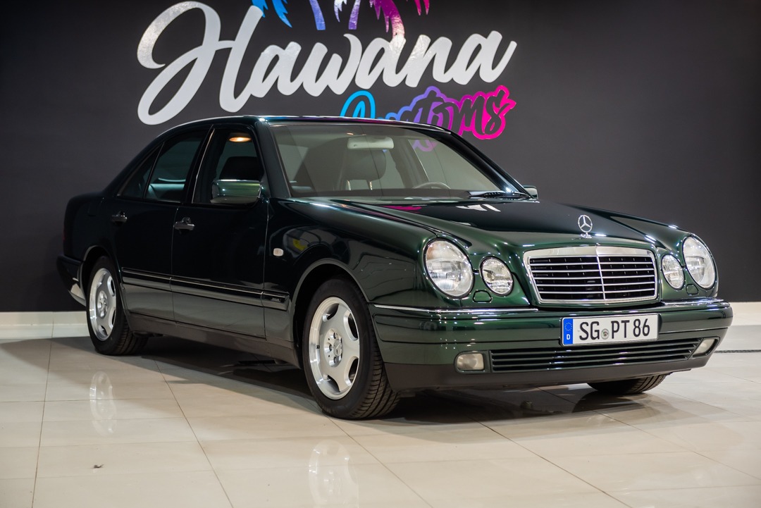 Zielony Mercedes-Benz W210 na tle ściany z logo 'Hawana Customs', widoczne detale chromowane i aluminiowe felgi.