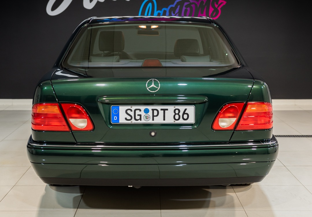 Tył ciemnozielonego Mercedesa klasy E (W210) z niemiecką tablicą rejestracyjną SG-PT 86, widoczny emblemat Mercedesa na klapie bagażnika, w tle czarna ściana z neonowym napisem.