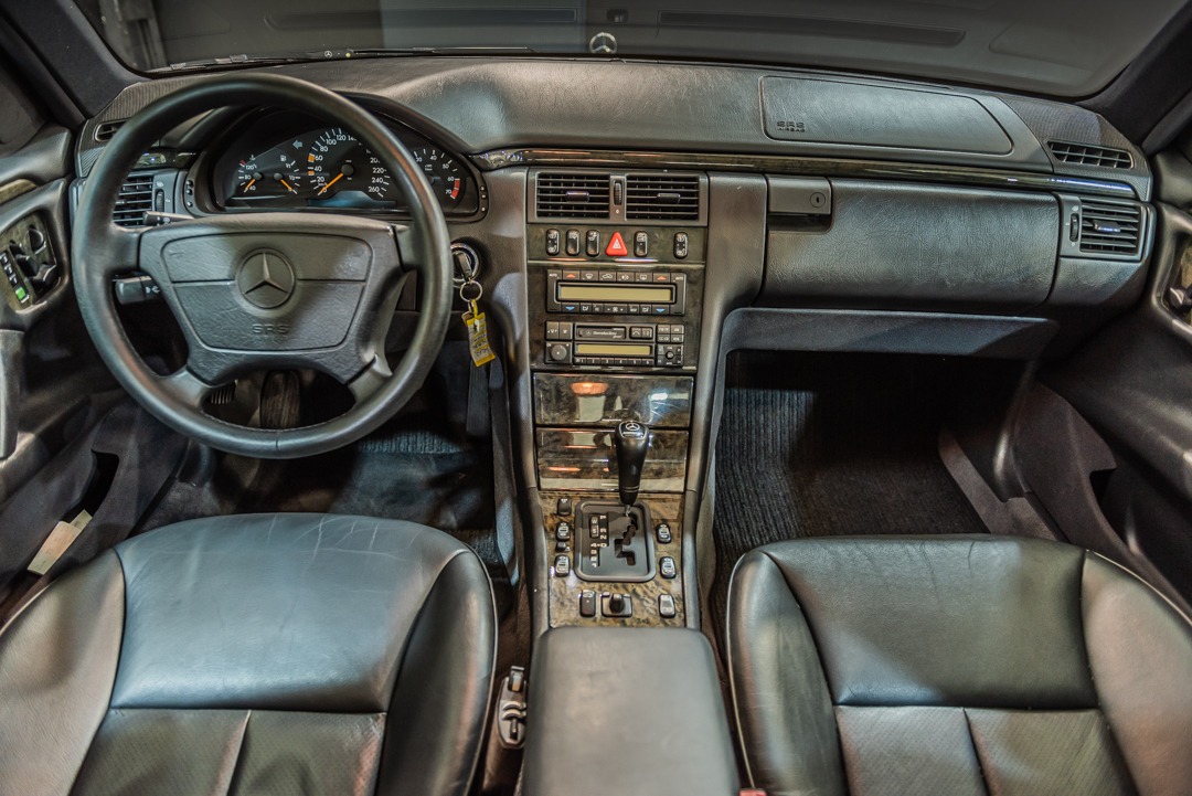 Wnętrze klasycznego Mercedesa W210 z czarną skórzaną tapicerką, widok na deskę rozdzielczą, kierownicę z logo, automatyczną skrzynię biegów i elementy wykończenia z drewna.