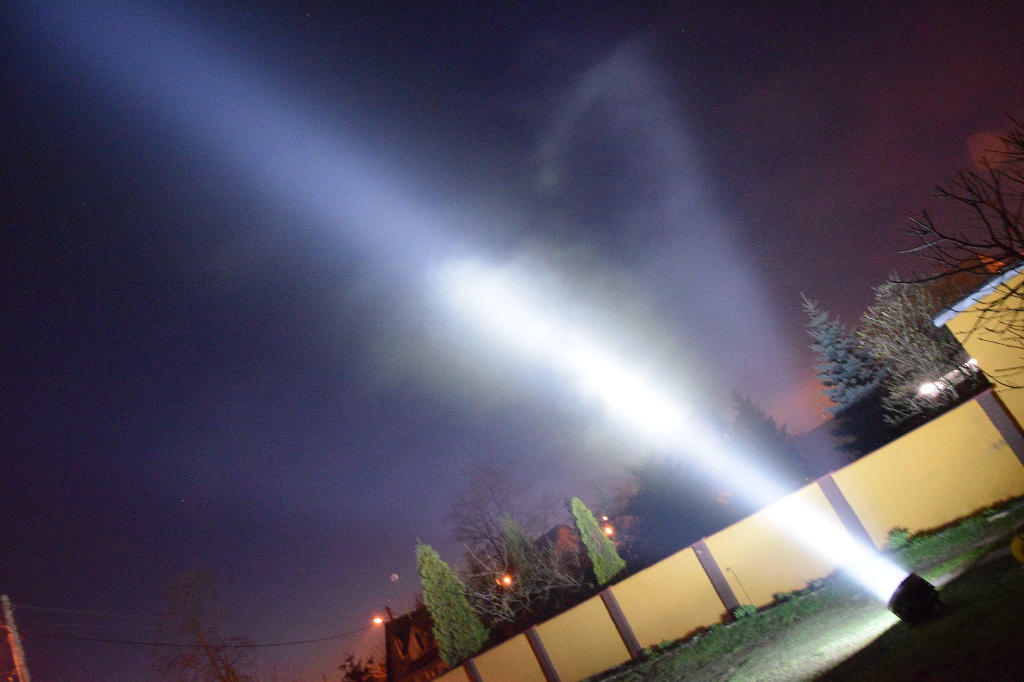 Oświetlenie elewacji domu reflektorem podczas nocnych prac w ogrodzie, widoczny snop światła i żółty mur.