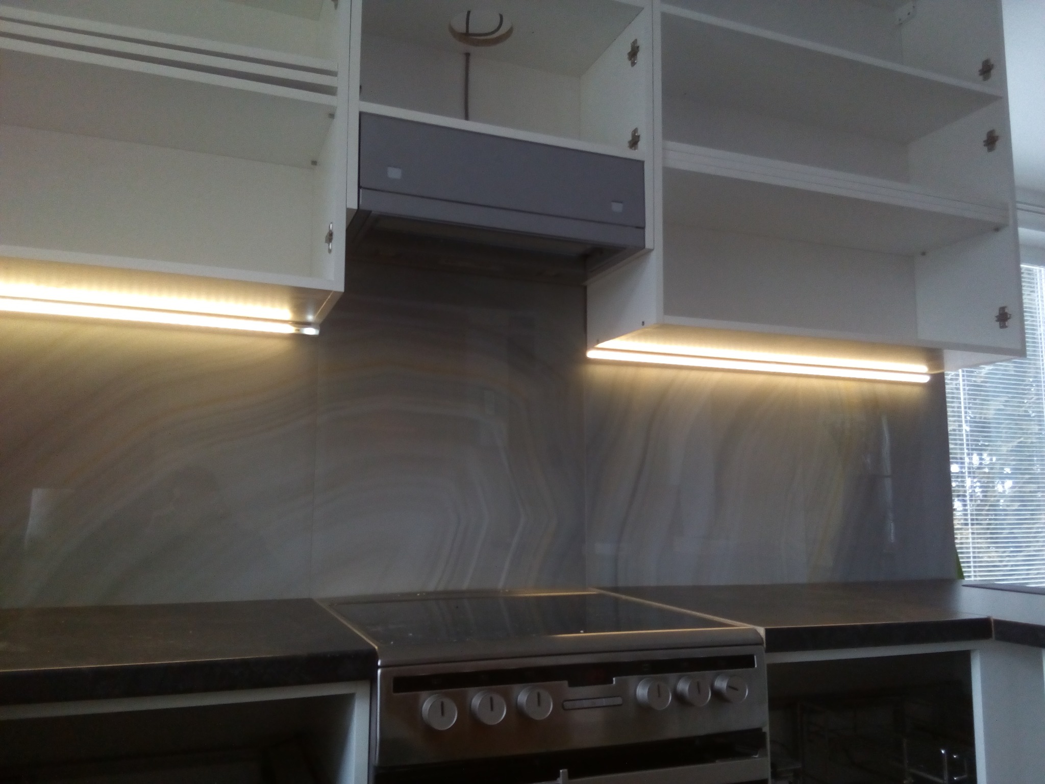 Kuchnia z białymi szafkami wiszącymi, podświetleniem LED pod szafkami, szarą płytą indukcyjną i okapem, marmurowy wzór na ścianie.