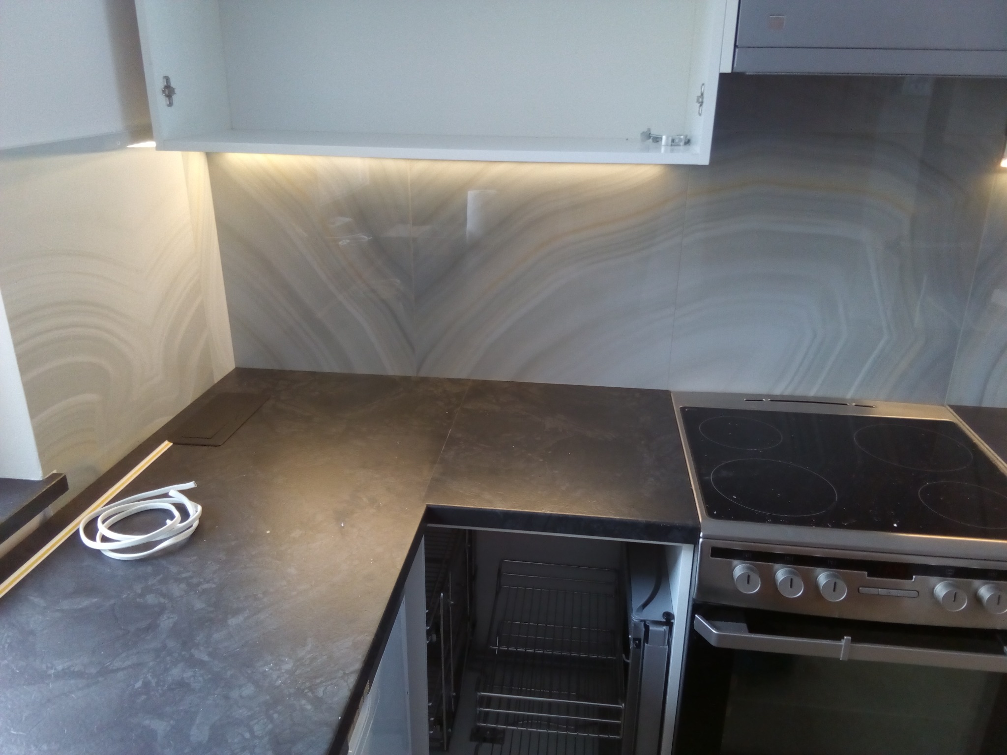 Nowoczesna kuchnia z czarnym blatem, marmurowymi płytkami na ścianie i oświetleniem LED pod szafkami wiszącymi; widoczny piekarnik z wysuwanym koszem oraz płyta indukcyjna.