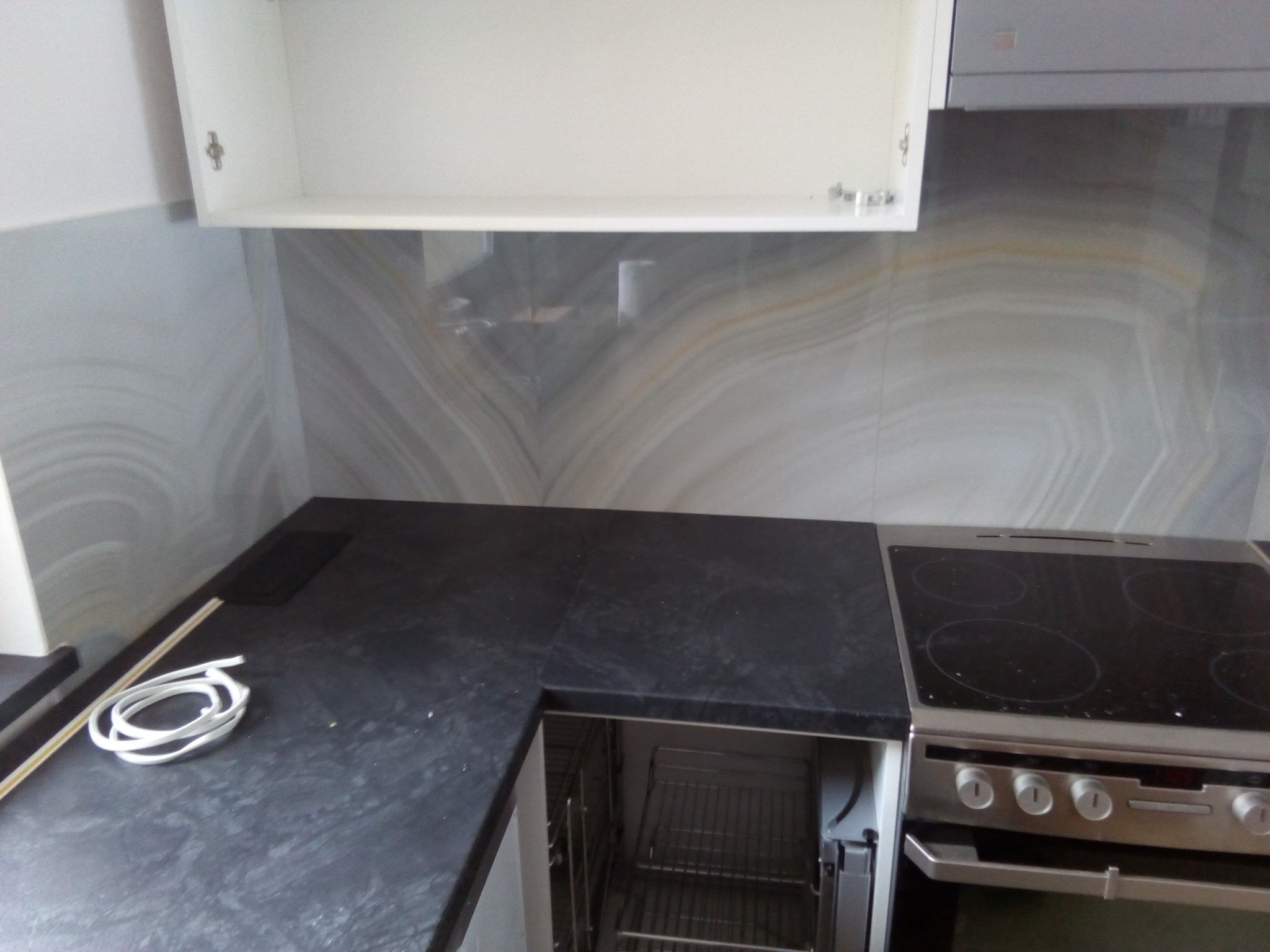 Fragment kuchni z czarnym blatem, płytą indukcyjną i szafką wiszącą na tle marmurowej ściany. Widoczny zwój kabla na blacie.