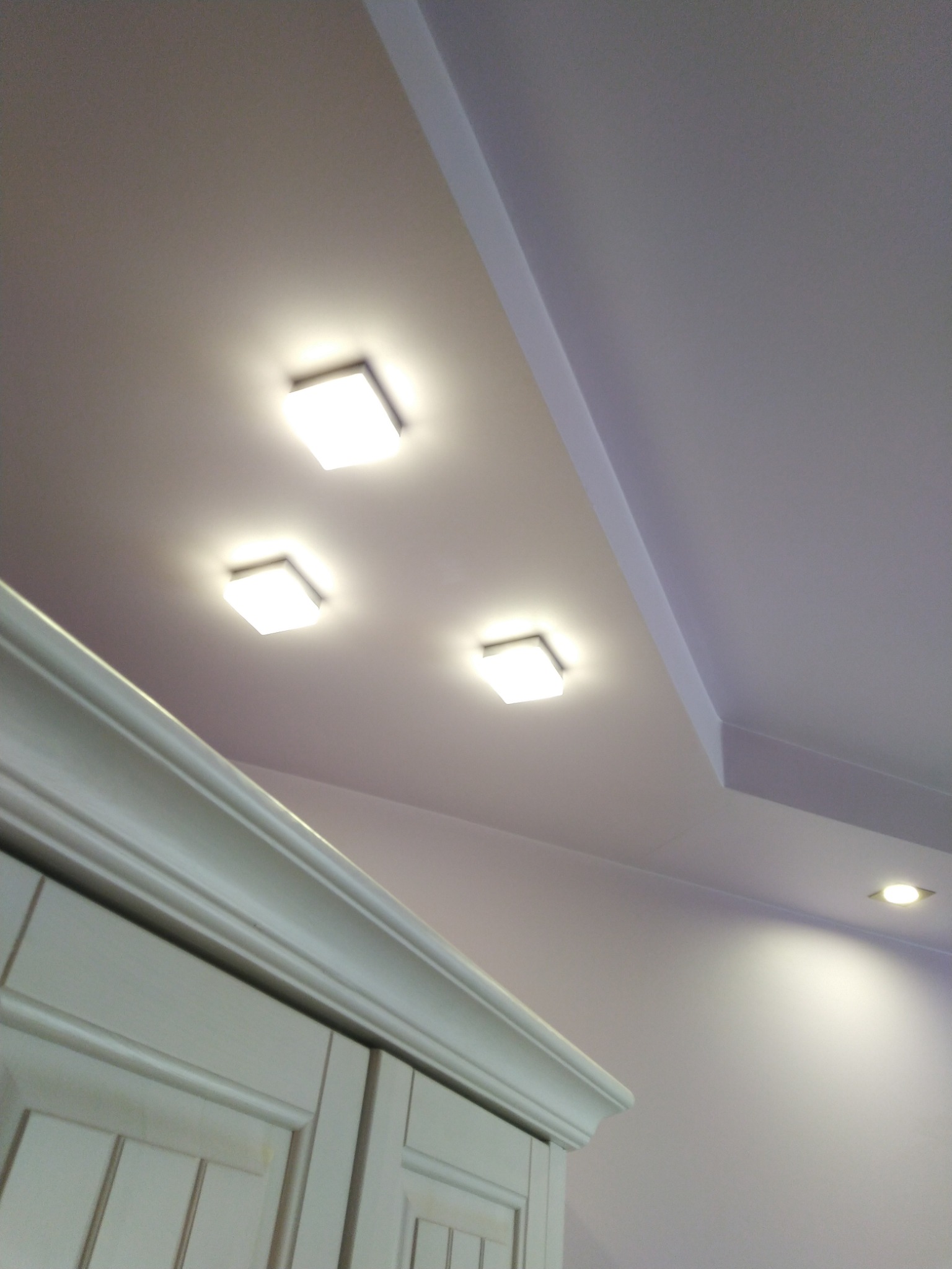 Biały sufit z trzema kwadratowymi lampami LED i jedną okrągłą, fragment białej szafki z dekoracyjnym frezowaniem na pierwszym planie.