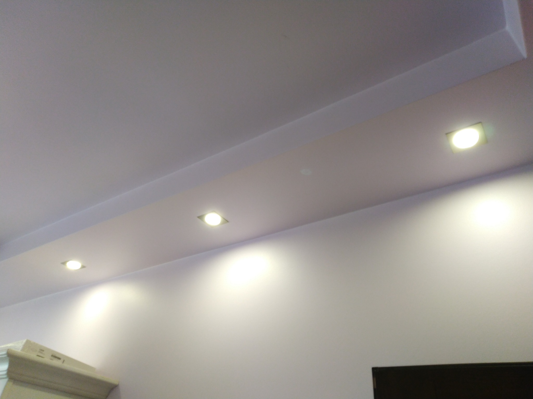 Podwieszany sufit z czterema kwadratowymi lampami LED, skierowanymi w dół, oświetlającymi ścianę w odcieniach bieli i fioletu.