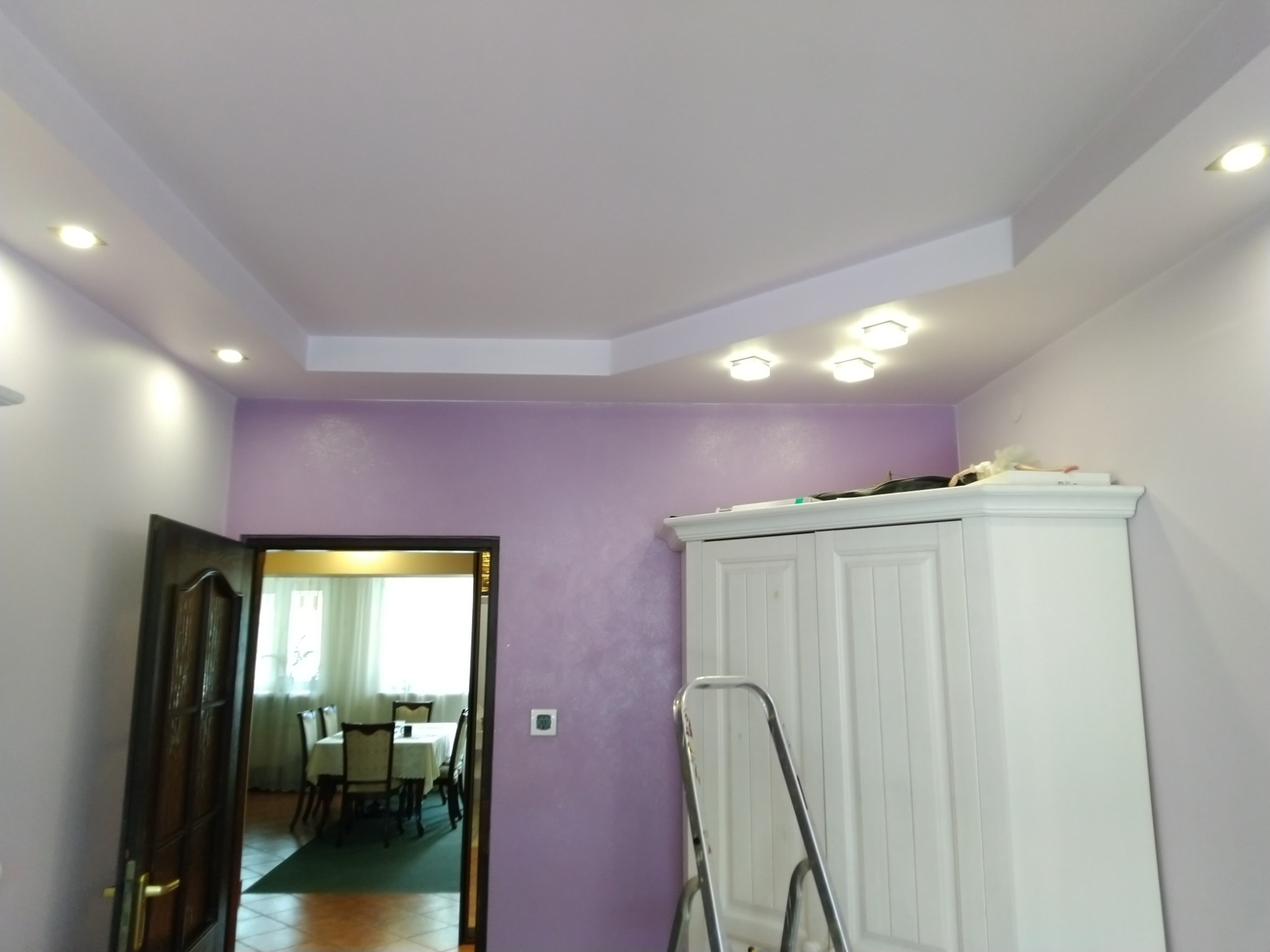 Fioletowa ściana z dekoracyjnym wykończeniem, biały sufit z wbudowanym oświetleniem, widoczny fragment szafy i drabiny, w tle otwarte drzwi do jadalni.