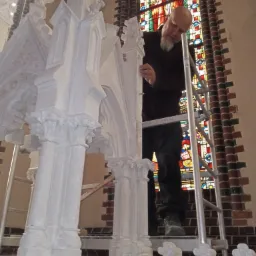 Pracownik na drabinie, kontrolujący fragment odrestaurowanej, bogato zdobionej, białej konstrukcji architektonicznej w kościele, z witrażem w tle.