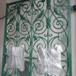 Świeżo pomalowana, zielona, ozdobna krata metalowa z motywem roślinnym, zabezpieczona folią przed uszkodzeniami, wisząca na ścianie.
