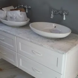 Łazienkowa szafka z szufladami i blatem z białego kamienia, na którym stoją dwie owalne, białe umywalki nablatowe. Jedna umywalka z opakowaniem producenta w środku.