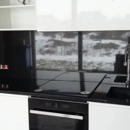 Czarny, błyszczący blat i panel kuchenny z kamienia z widocznymi gniazdkami i nowoczesną baterią zlewozmywakową na tle okna z zimowym krajobrazem.