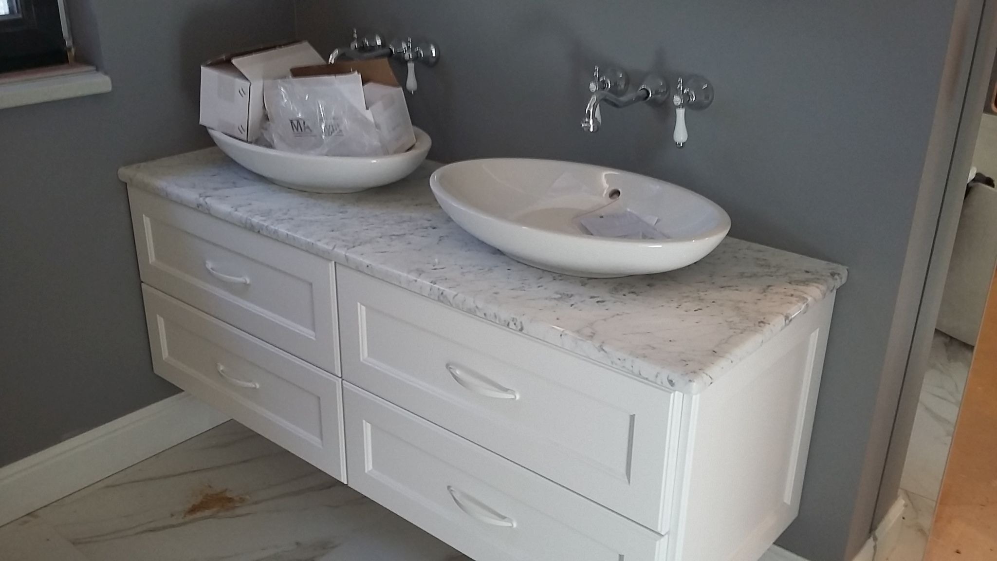 Łazienkowa szafka z szufladami i blatem z białego kamienia, na którym stoją dwie owalne, białe umywalki nablatowe. Jedna umywalka z opakowaniem producenta w środku.