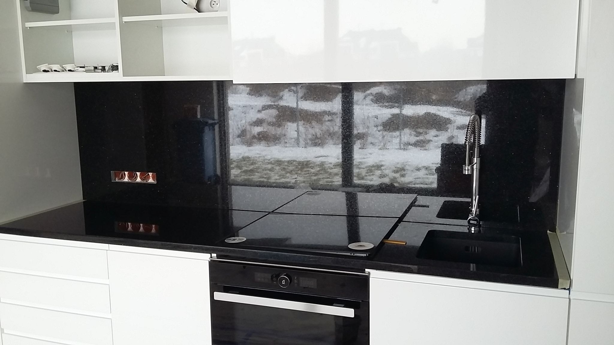 Czarny, błyszczący blat i panel kuchenny z kamienia z widocznymi gniazdkami i nowoczesną baterią zlewozmywakową na tle okna z zimowym krajobrazem.