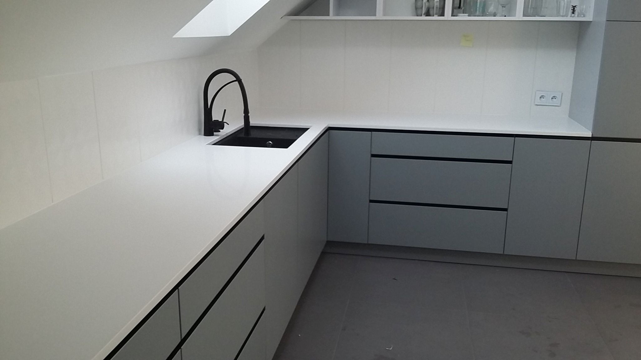 Biały, narożny blat kamienny z czarnym zlewem i baterią, w nowoczesnej kuchni na poddaszu ze szarymi szafkami i płytkami ściennymi.