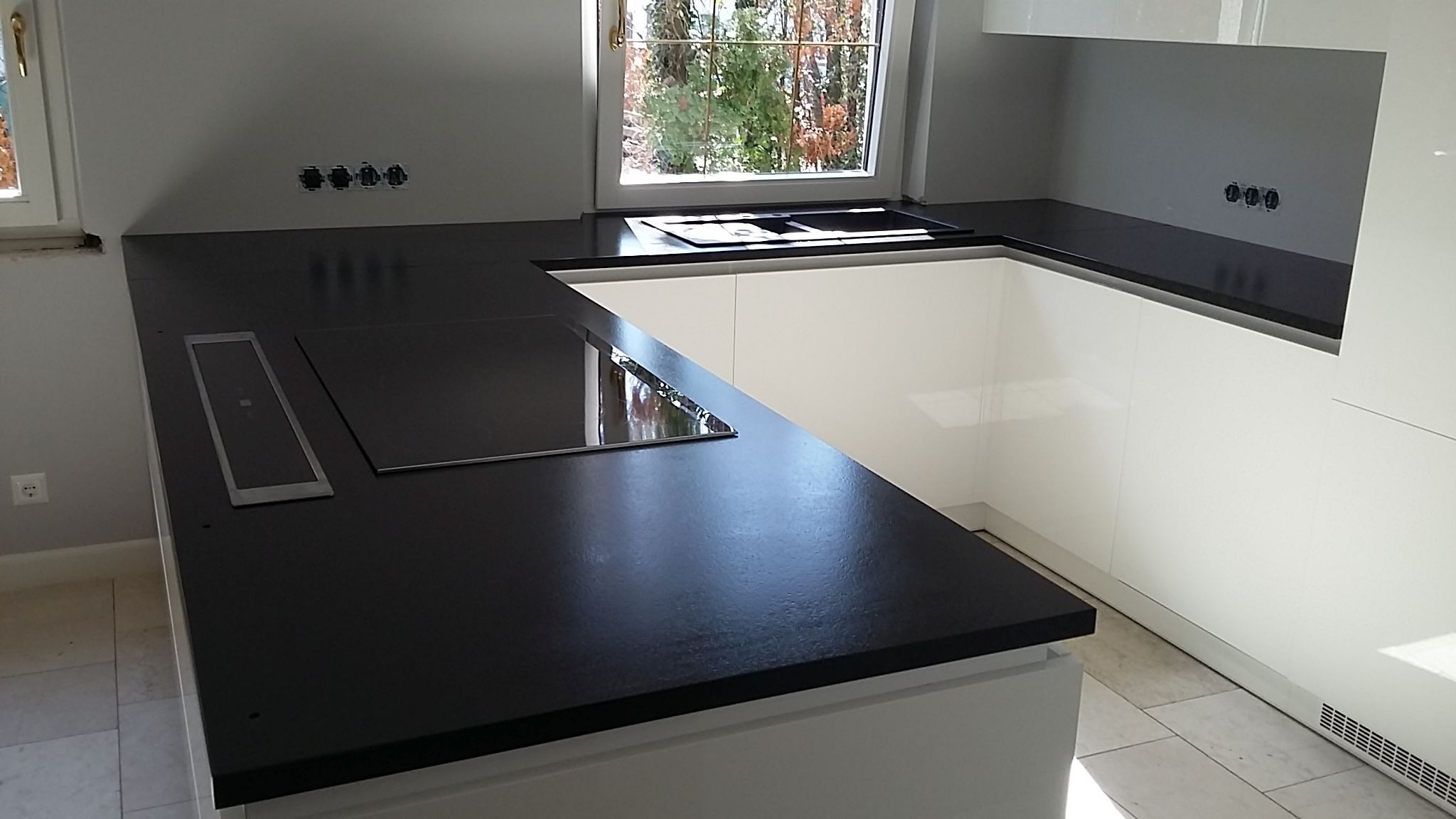 Czarny blat kuchenny z wbudowaną płytą indukcyjną i gniazdem elektrycznym, widok na okno z zielenią w tle, białe szafki kuchenne w nowoczesnym stylu.