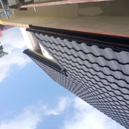 Widok z dołu na fragment dachu pokrytego szarą blachodachówką z czarnymi rynnami, na tle błękitnego nieba z chmurami i fragmentami zieleni drzew