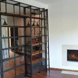 Metalowa konstrukcja regału z drewnianymi półkami i szklanymi drzwiami oddzielająca przestrzeń w salonie z kominkiem, widoczne schody w tle.