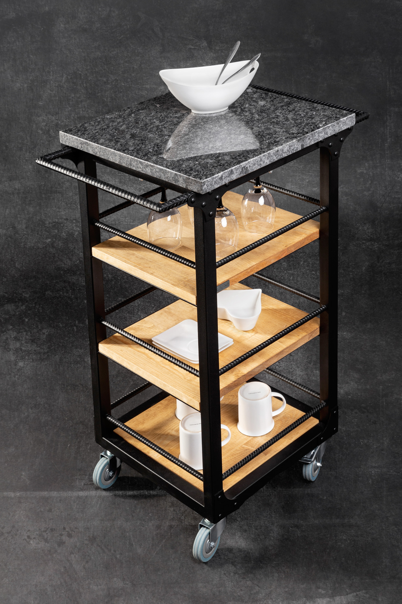 Wózek kuchenny na kółkach z blatem z ciemnego granitu, drewnianymi półkami i czarną metalową konstrukcją, na którym znajdują się naczynia: miska, szklanki, talerze i kubki.