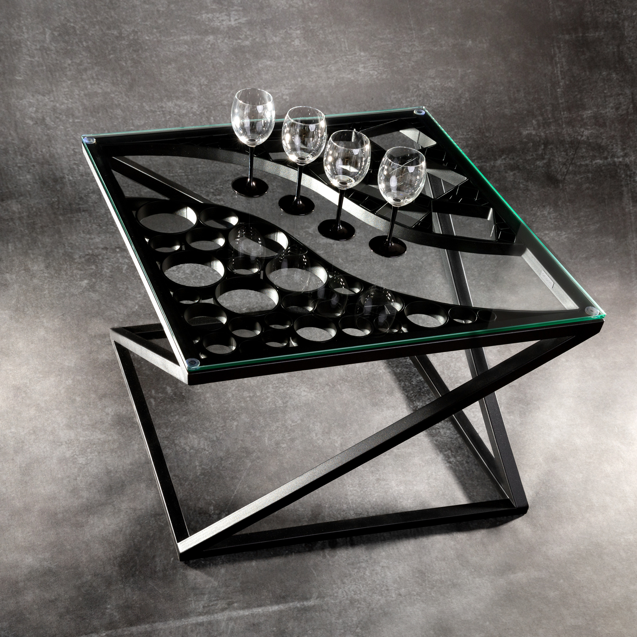 Nowoczesny stolik kawowy z czarną metalową podstawą i szklanym blatem z otworami na kieliszki, na którym stoją cztery kieliszki do wina, na tle szarej ściany.