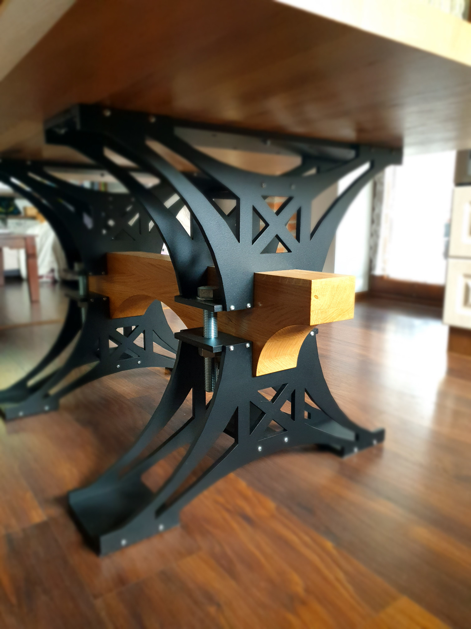 Stół z blatem z jasnego drewna i metalową, czarną konstrukcją nóg z dekoracyjnymi wycięciami, połączoną drewnianymi elementami i śrubami.