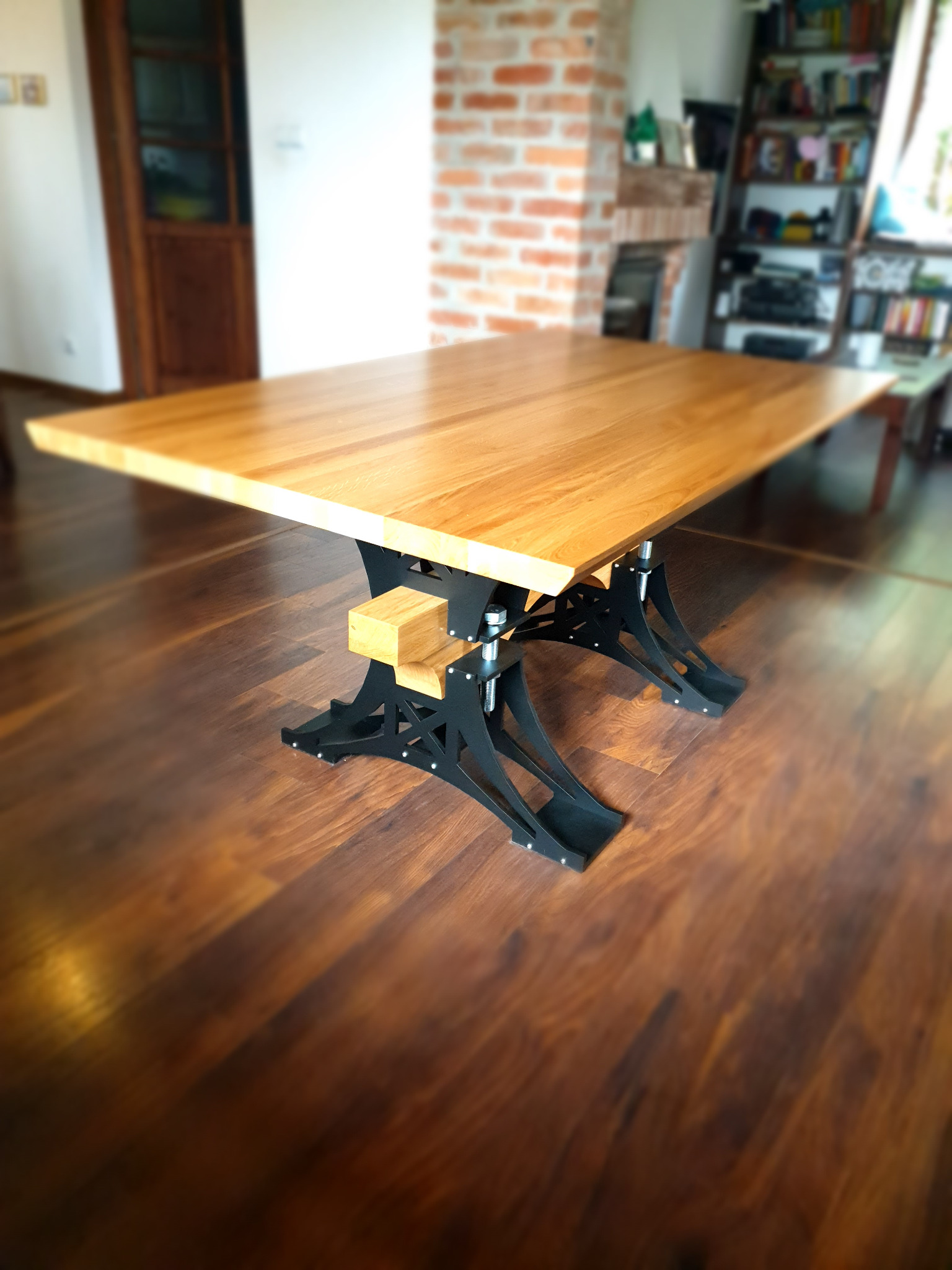 Stół z blatem z jasnego drewna i czarnymi, metalowymi nogami o regulowanej wysokości, na drewnianej podłodze w tle ściana z cegły i regał z książkami.