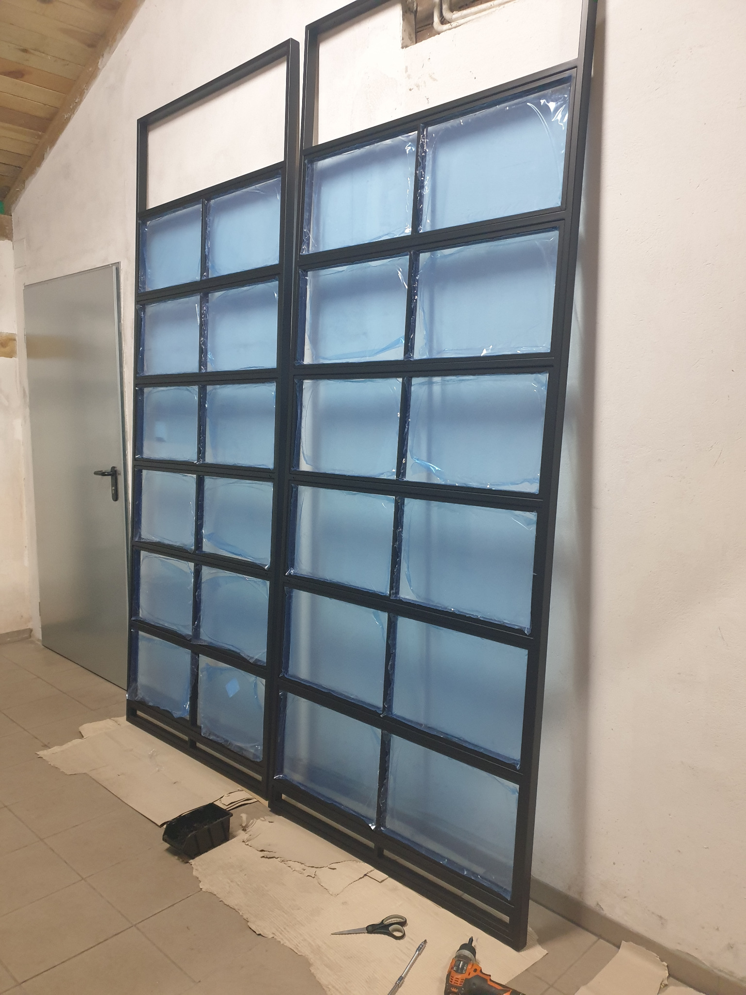 Dwa czarne, metalowe ramy z szybami zabezpieczonymi folią, ustawione obok siebie, w tle biała ściana i szare drzwi, na podłodze karton i narzędzia.