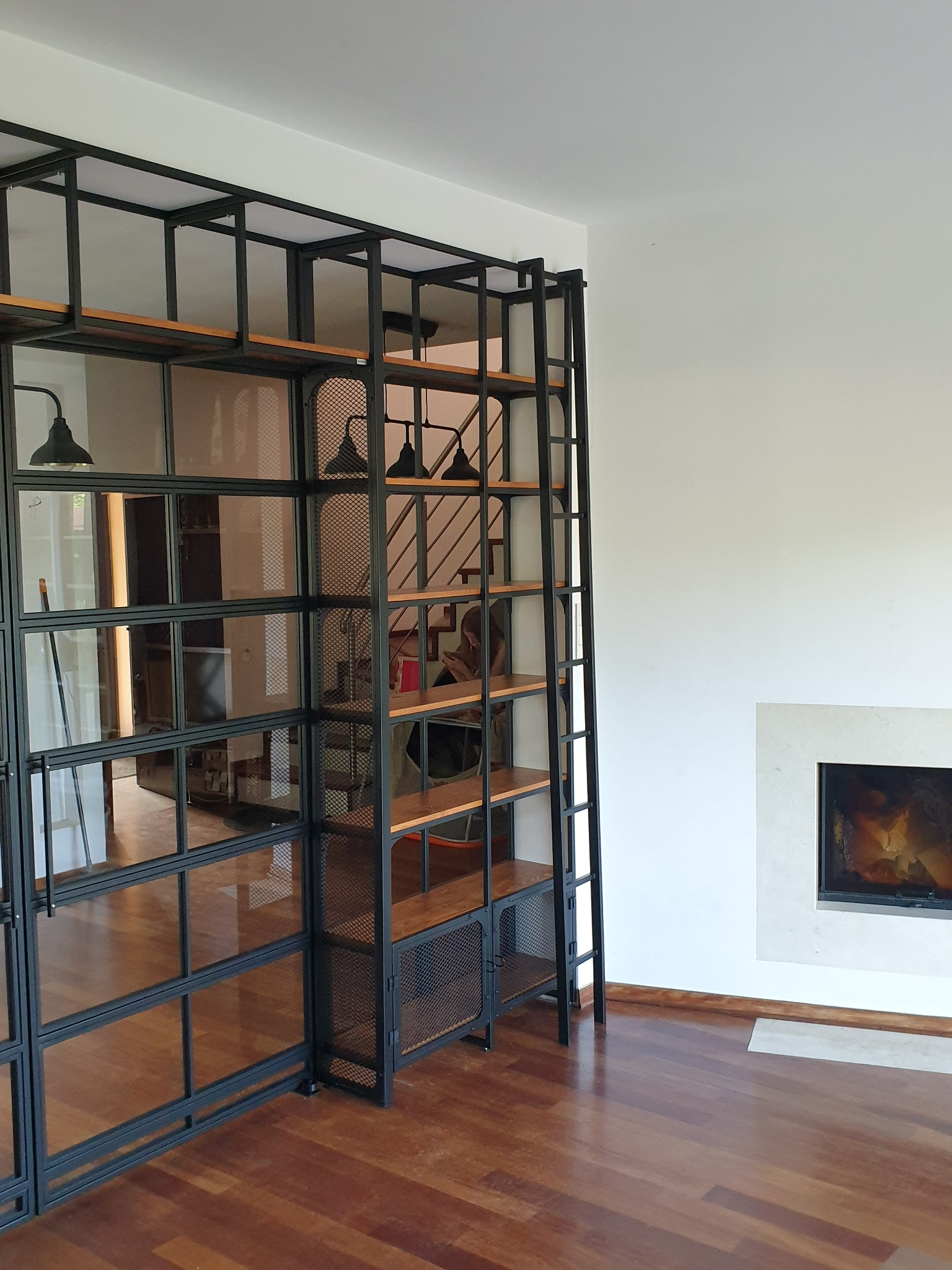 Metalowa konstrukcja regału z drewnianymi półkami i szklanymi drzwiami oddzielająca przestrzeń w salonie z kominkiem, widoczne schody w tle.