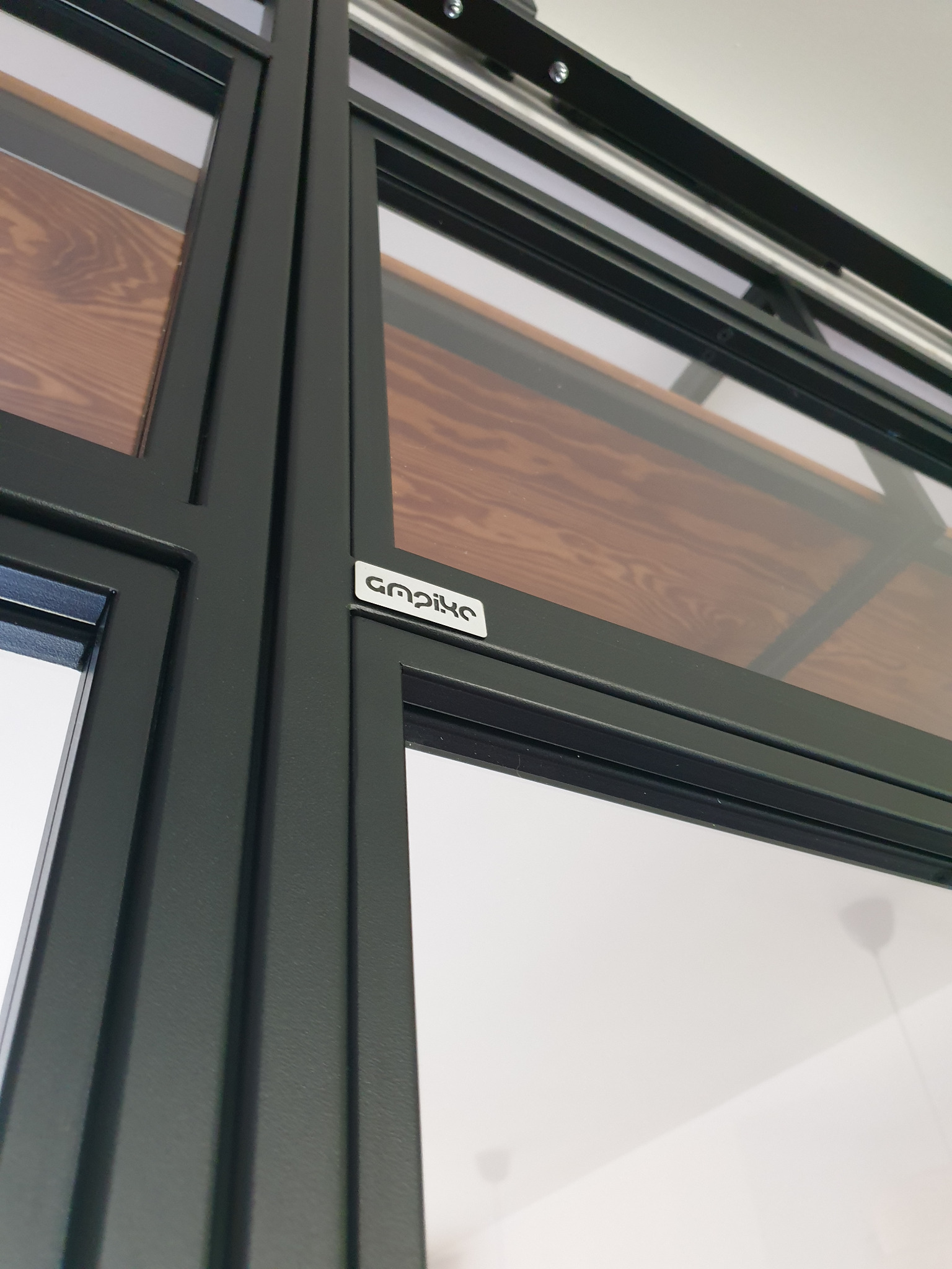 Szczegółowe ujęcie czarnej ramy drzwi przesuwnych z drewnianym panelem, widoczny system szyn i logo producenta.