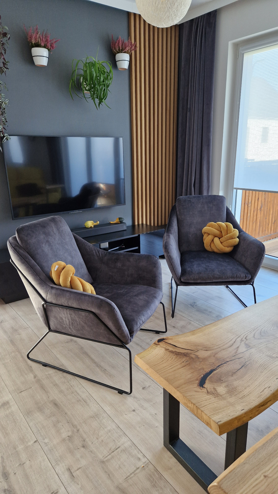 Nowoczesny salon z dwoma grafitowymi fotelami na metalowych nogach, drewnianym stolikiem kawowym z naturalną krawędzią, telewizorem na ścianie i dekoracyjnymi panelami pionowymi z drewna.