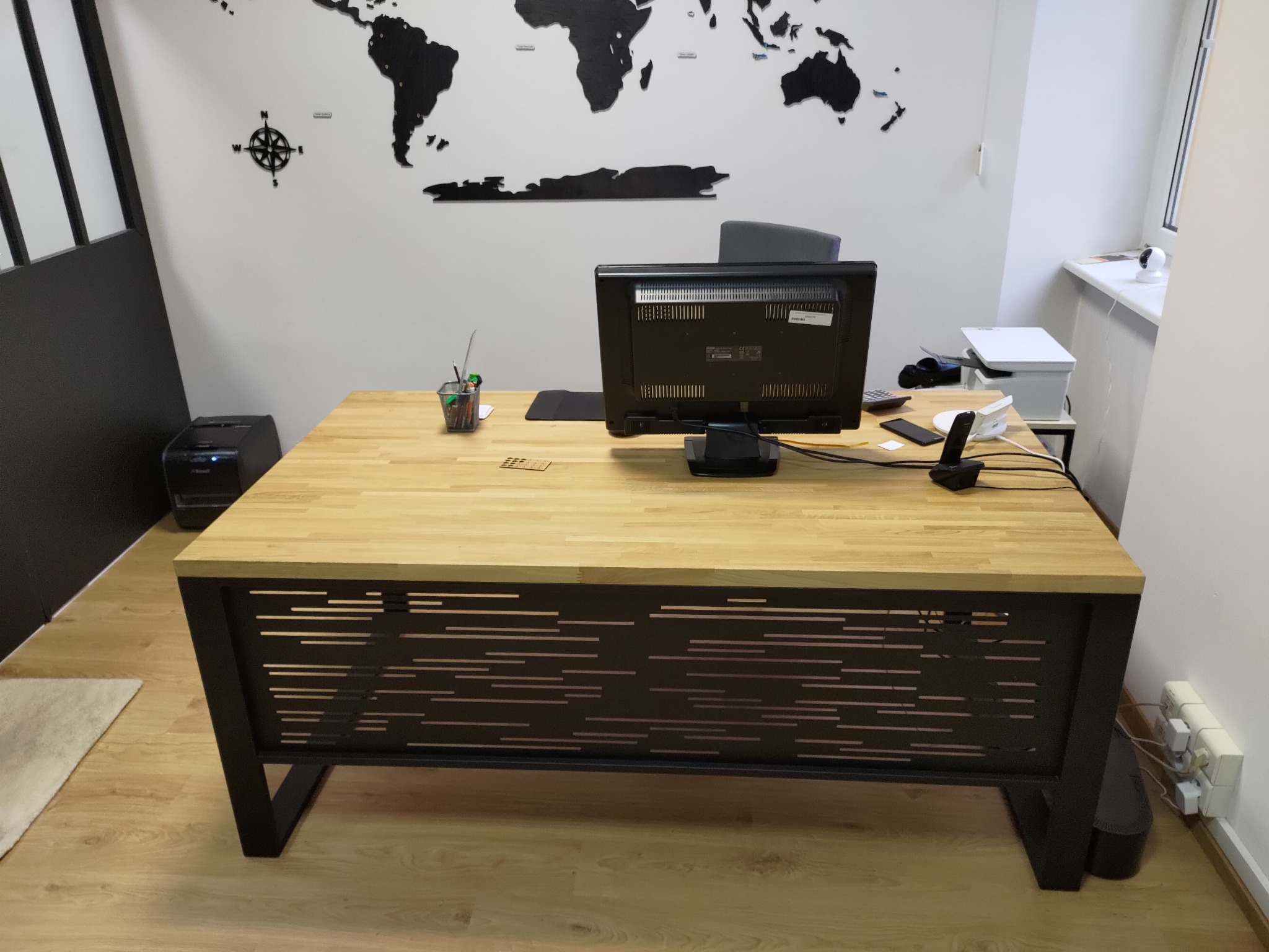 Biurko z blatem z jasnego drewna i metalową podstawą z poziomymi wycięciami, na tle ściany z mapą świata, monitor, przybory biurowe i drukarka w tle.