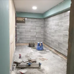 Wnętrze w trakcie prac wykończeniowych, ściany częściowo pokryte szarymi płytkami imitującymi drewno, widoczne narzędzia glazurnicze, wiadro z zaprawą i niebieski taboret.