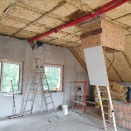 Ocieplone poddasze w Opalenicy w trakcie prac wykończeniowych, widoczne wełna mineralna, czerwone rury wentylacyjne pod sufitem, drabiny, okna i narzędzia.