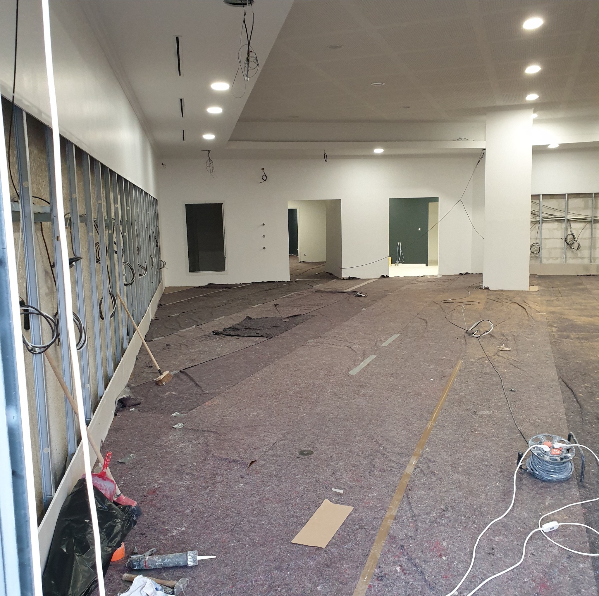 Wnętrze w trakcie generalnego remontu: widoczne metalowe profile na ścianie, zabezpieczona podłoga, przewody elektryczne zwisające z sufitu i narzędzia budowlane.
