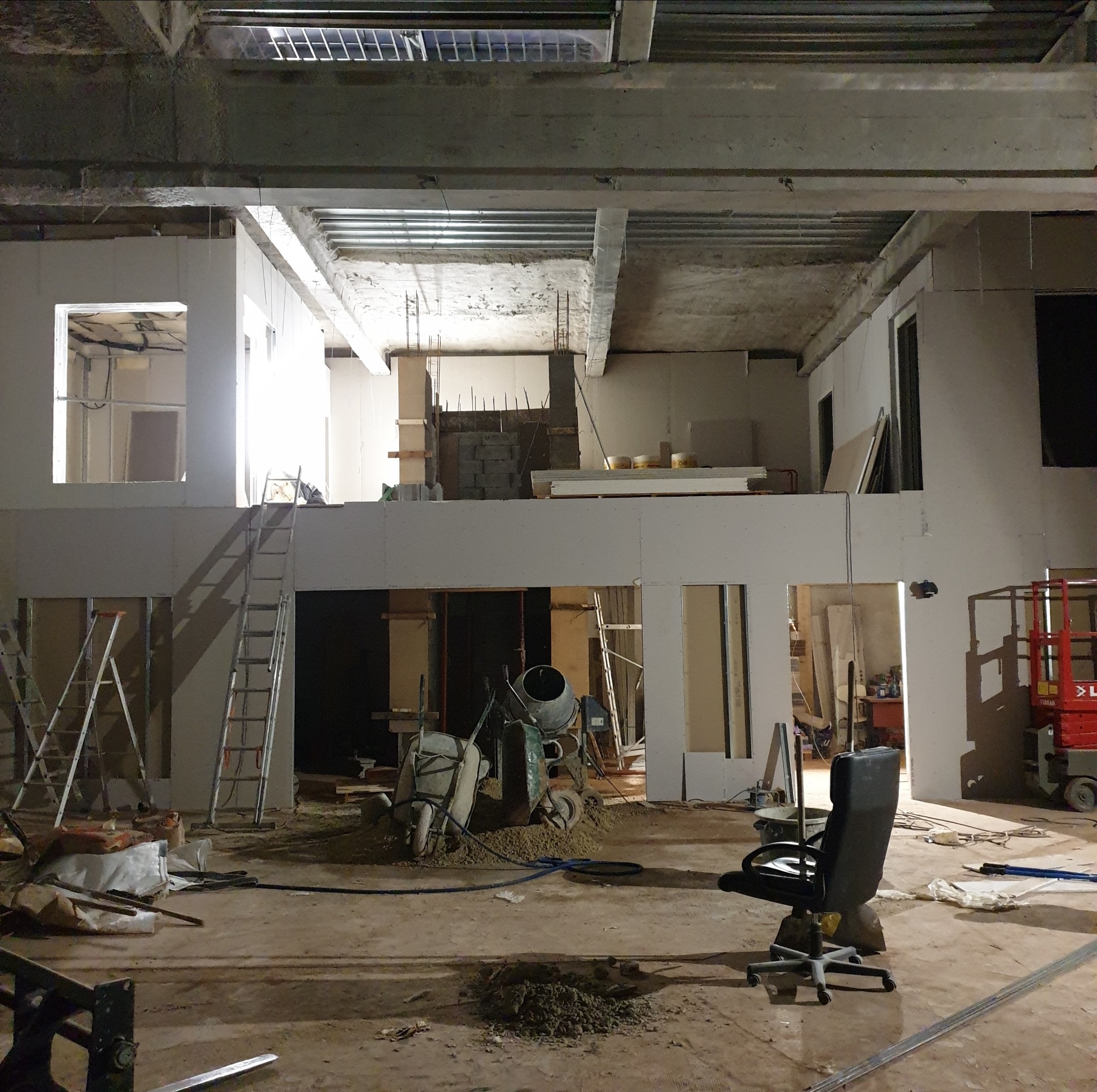 Wnętrze budynku w trakcie remontu, widoczne surowe ściany, drabiny, betoniarka, wózki budowlane, gruz i krzesło biurowe.
