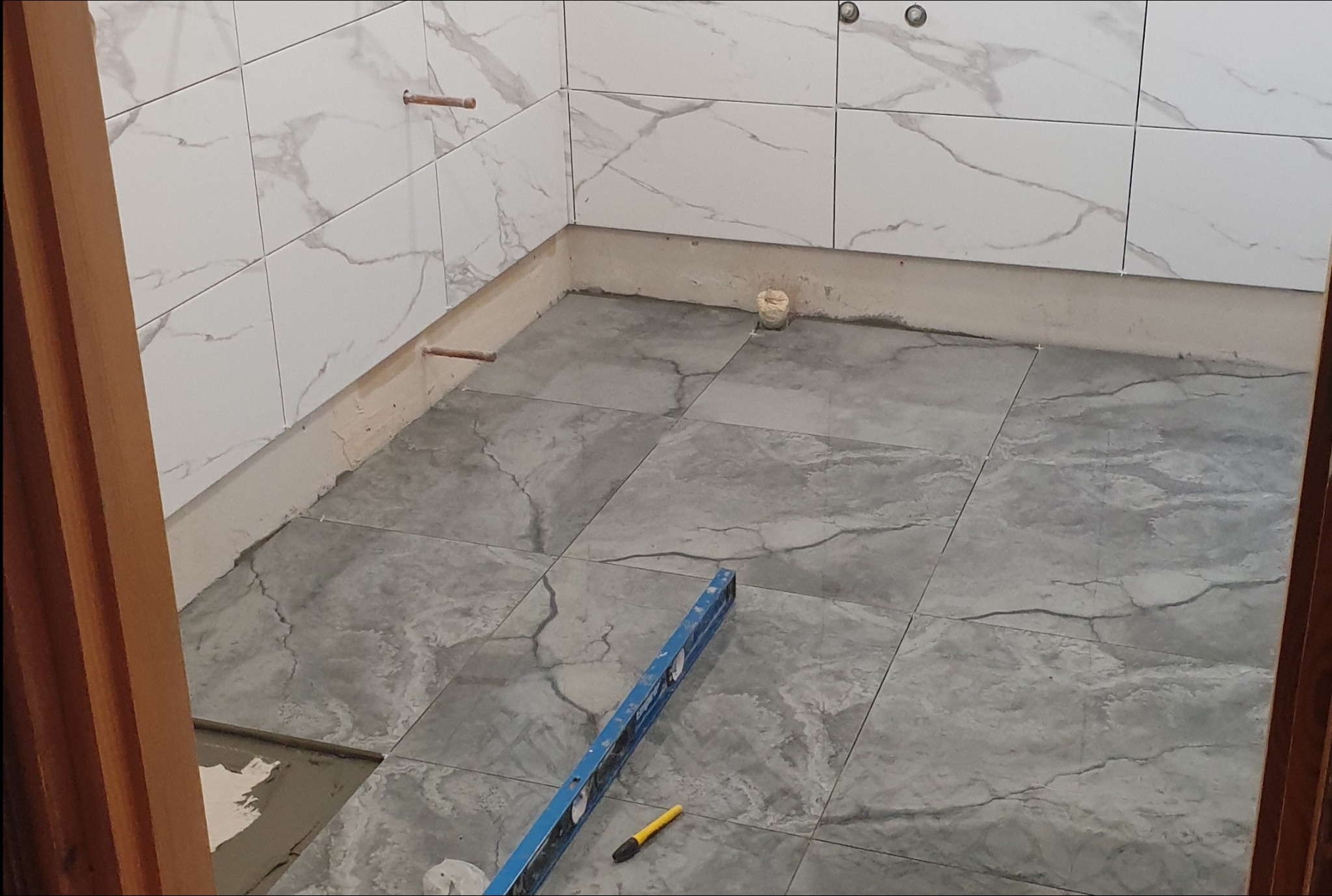 Podłoga w trakcie układania szarych płytek imitujących marmur, widoczna poziomica i ołówek glazurniczy, ściany wyłożone białymi płytkami z szarym wzorem marmuru, wystające rurki instalacji.