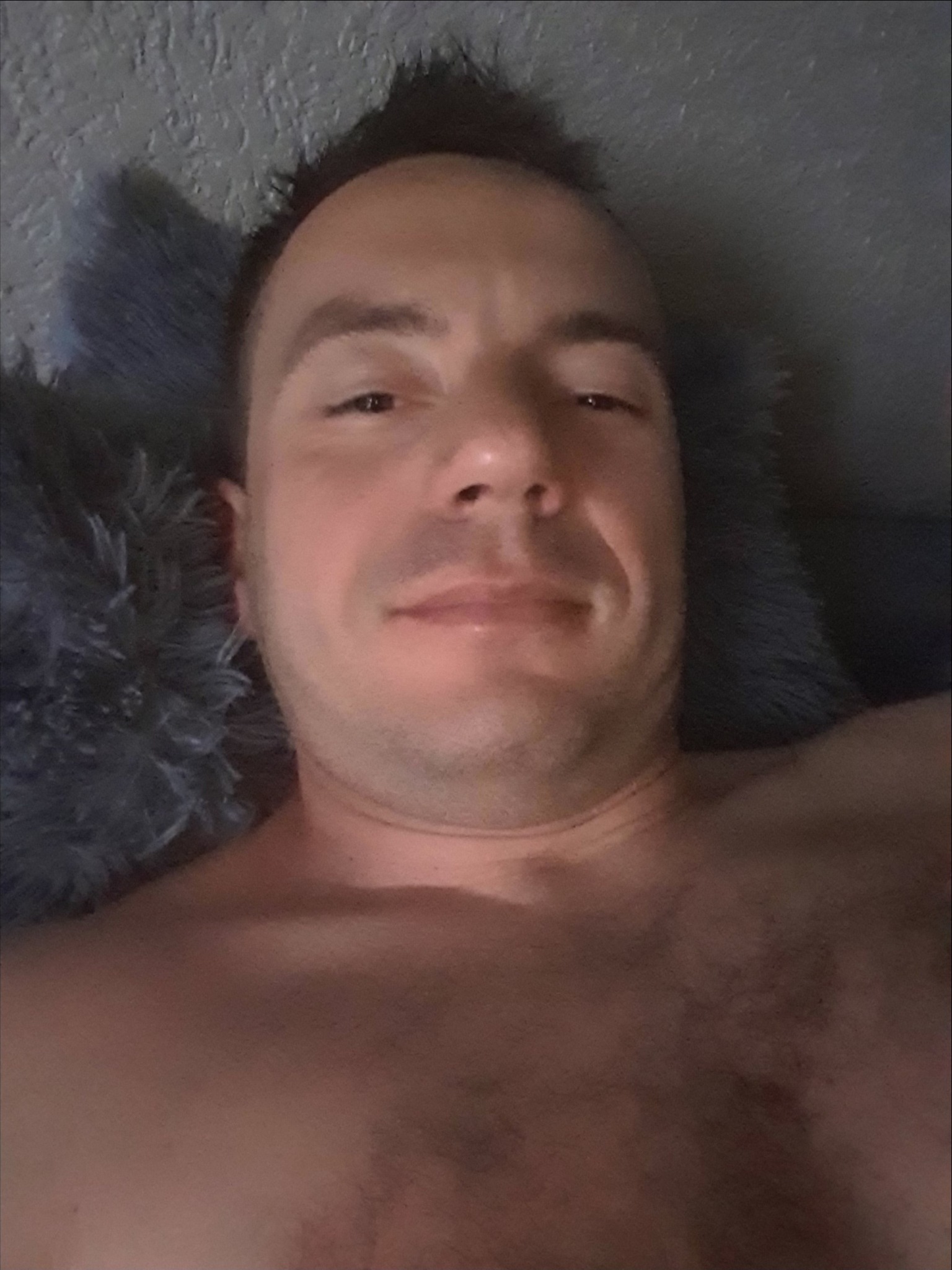 Portret mężczyzny leżącego na szarym, futrzanym dywanie, z widoczną górną częścią nagiego ciała.