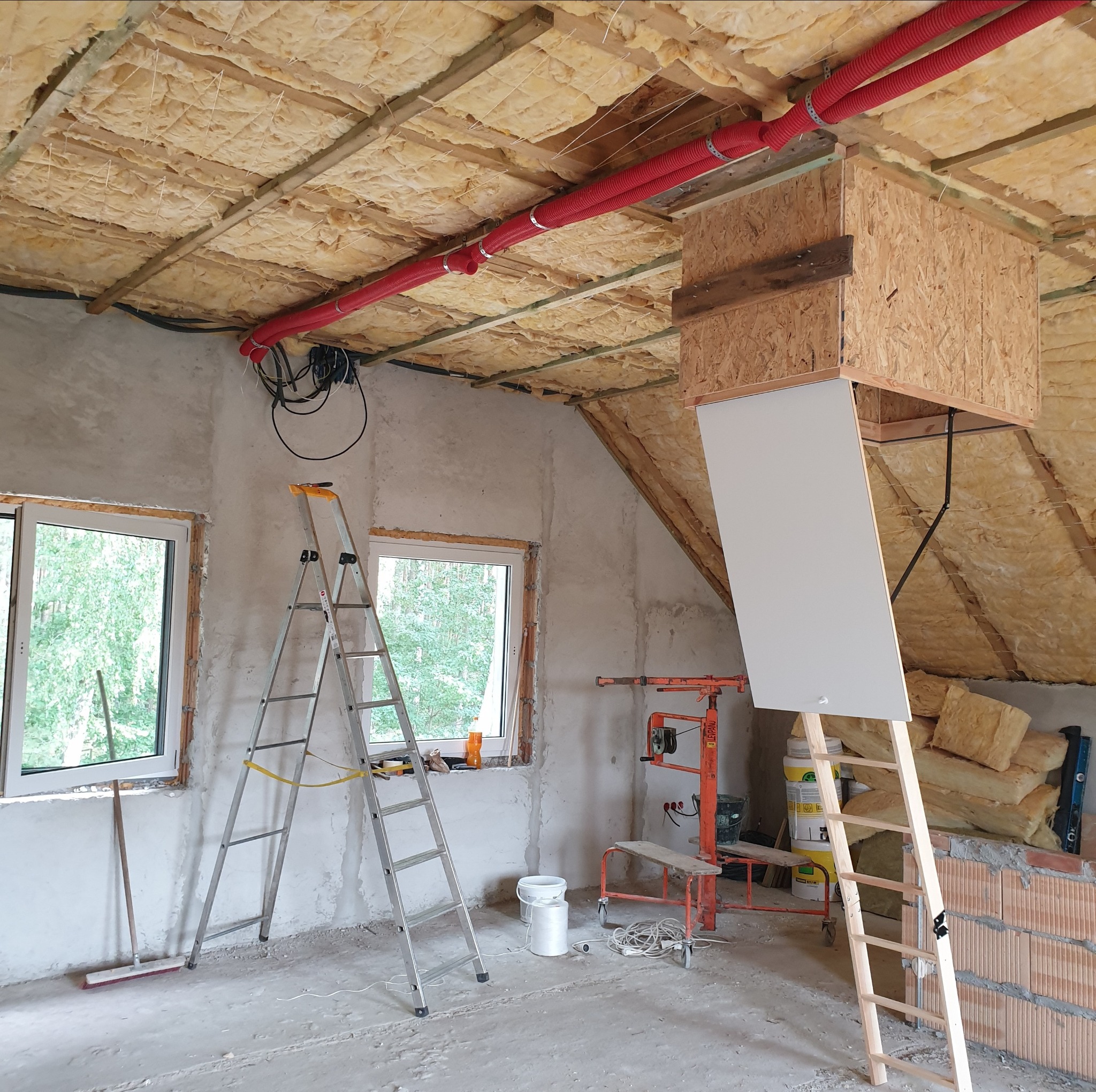 Ocieplone poddasze w Opalenicy w trakcie prac wykończeniowych, widoczne wełna mineralna, czerwone rury wentylacyjne pod sufitem, drabiny, okna i narzędzia.