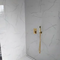 Nowoczesny prysznic walk-in z białymi płytkami imitującymi marmur i złotą armaturą, w tym deszczownica i bateria podtynkowa.