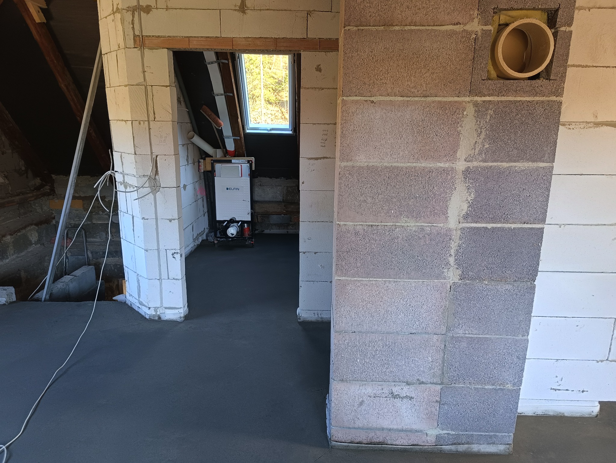 Wnętrze w trakcie budowy: wylana szara posadzka, ściany z bloczków betonowych, widoczny piec CO w głębi, fragment instalacji wentylacyjnej w ścianie.