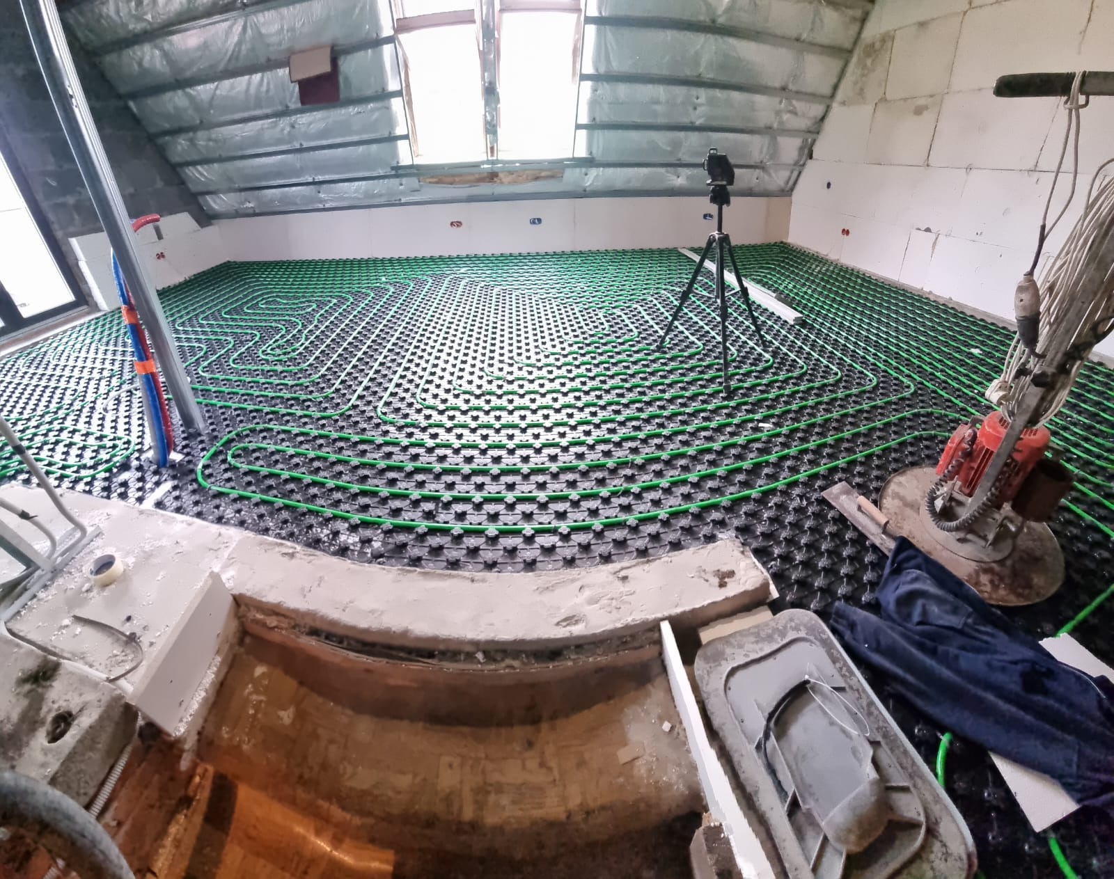 Układanie zielonych rur ogrzewania podłogowego na czarnej macie w poddaszu, widoczny szlifierka do posadzek i poziomica na trójnogu.