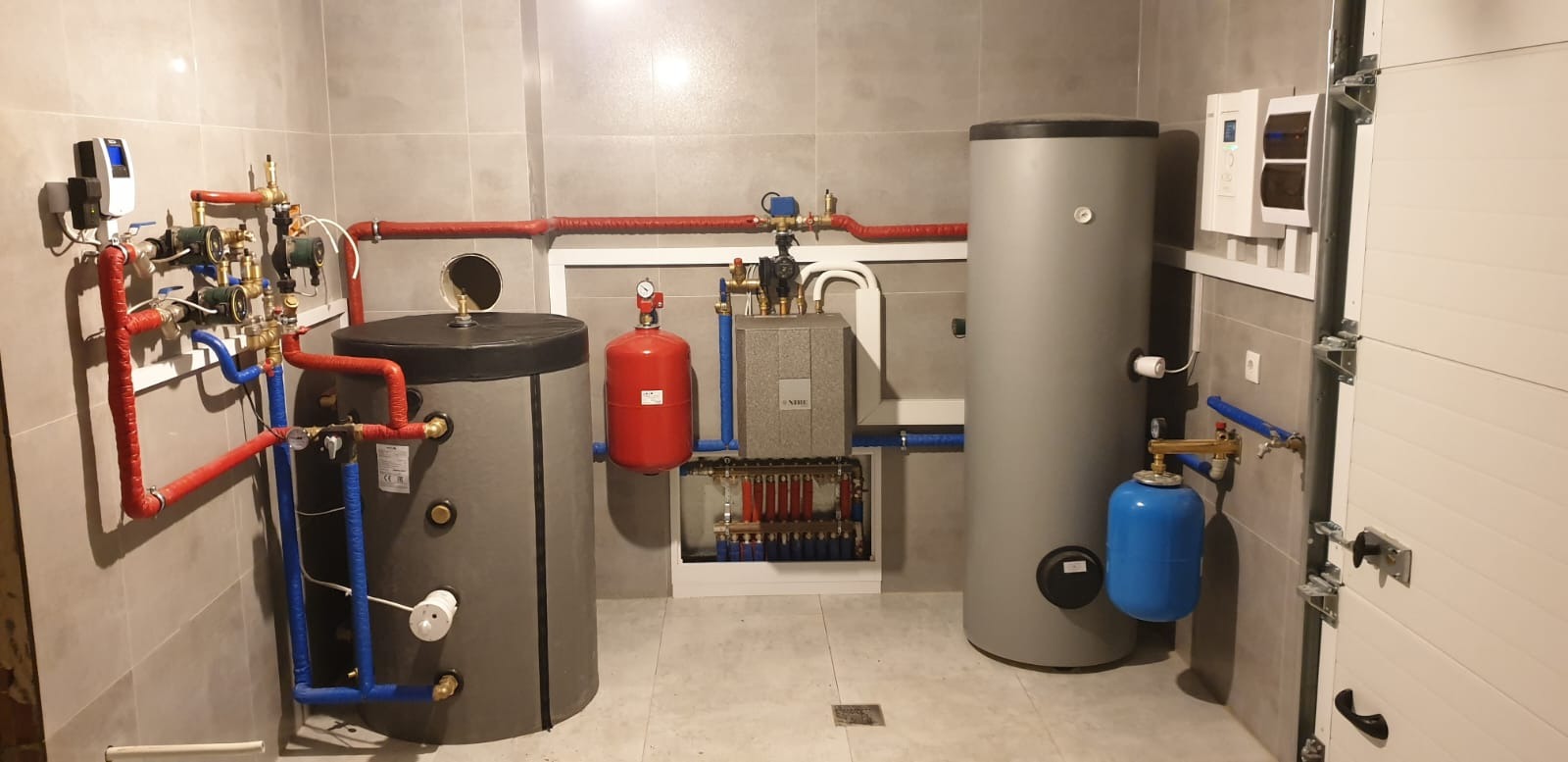 Kompletna instalacja grzewcza w kotłowni: dwa szare zbiorniki akumulacyjne, czerwone i niebieskie rury, rozdzielacz podłogówki w zabudowie, biały sterownik na ścianie z szarymi płytkami.