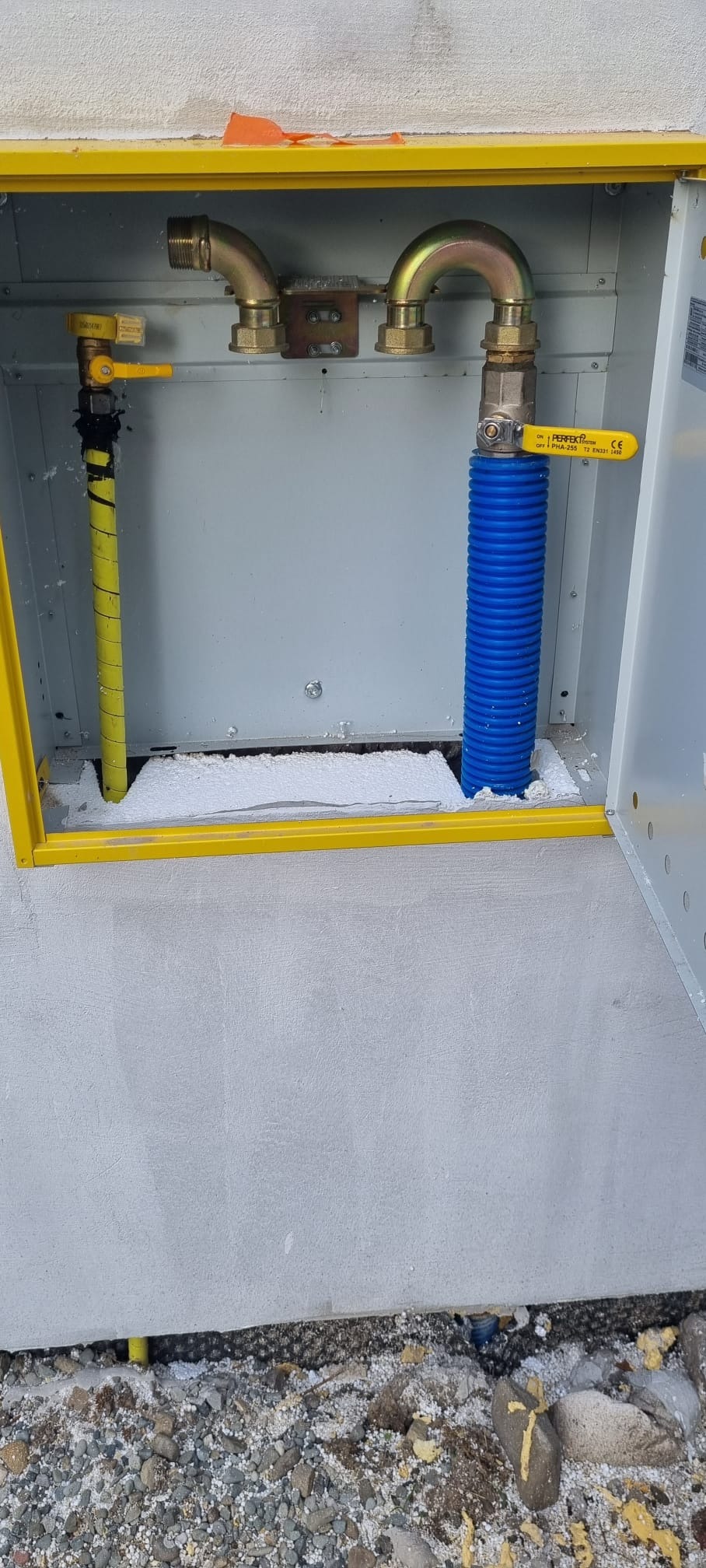 Przyłącze gazowe w żółtej skrzynce z widocznymi rurami: żółtą i niebieską, zabezpieczone pianką montażową, na tle szarego tynku i kamieni.