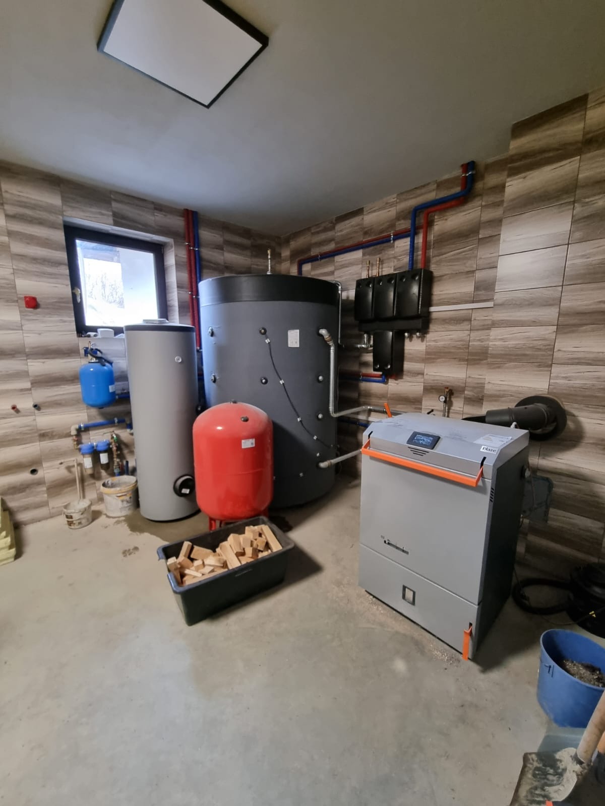Kotłownia z piecem, zbiornikami akumulacyjnymi, naczyniem wzbiorczym i drewnem opałowym w plastikowym pojemniku, widoczne rury instalacji grzewczej.