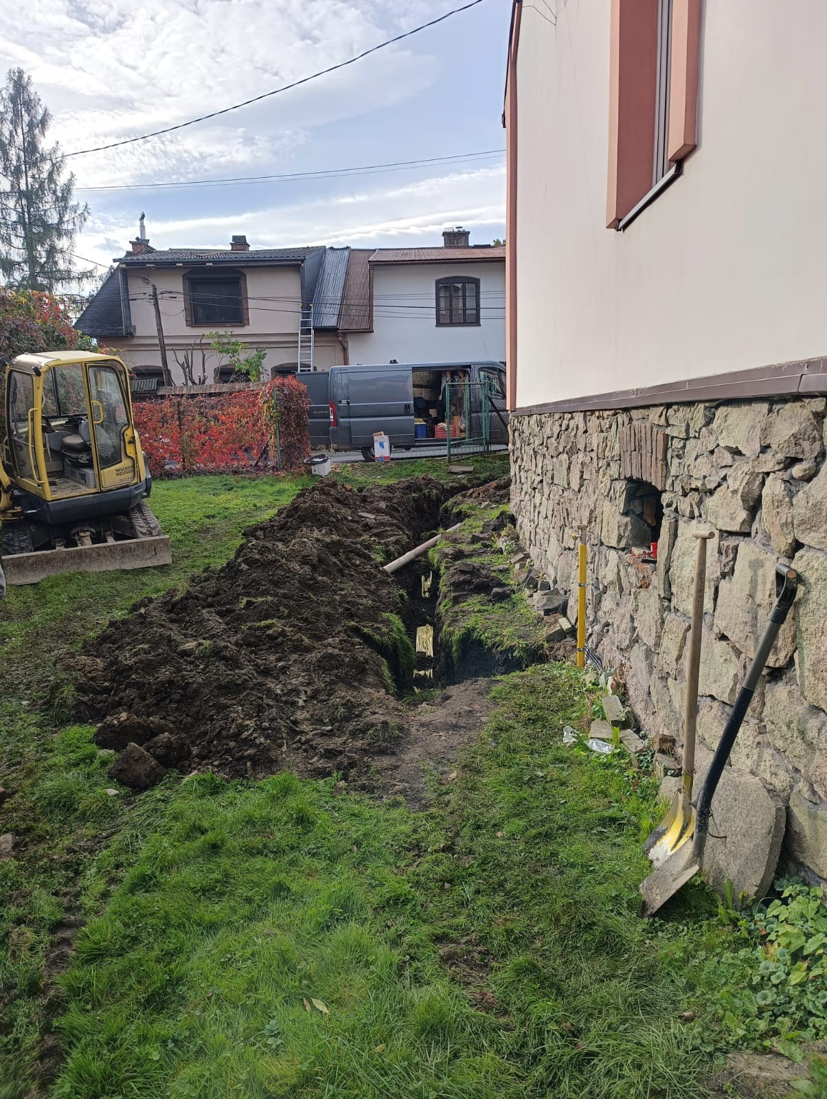 Wykop pod instalację w Osielcu, widoczny żółty mini-koparka, szpadle oparte o kamienną ścianę budynku, odkryte rury w wykopie.