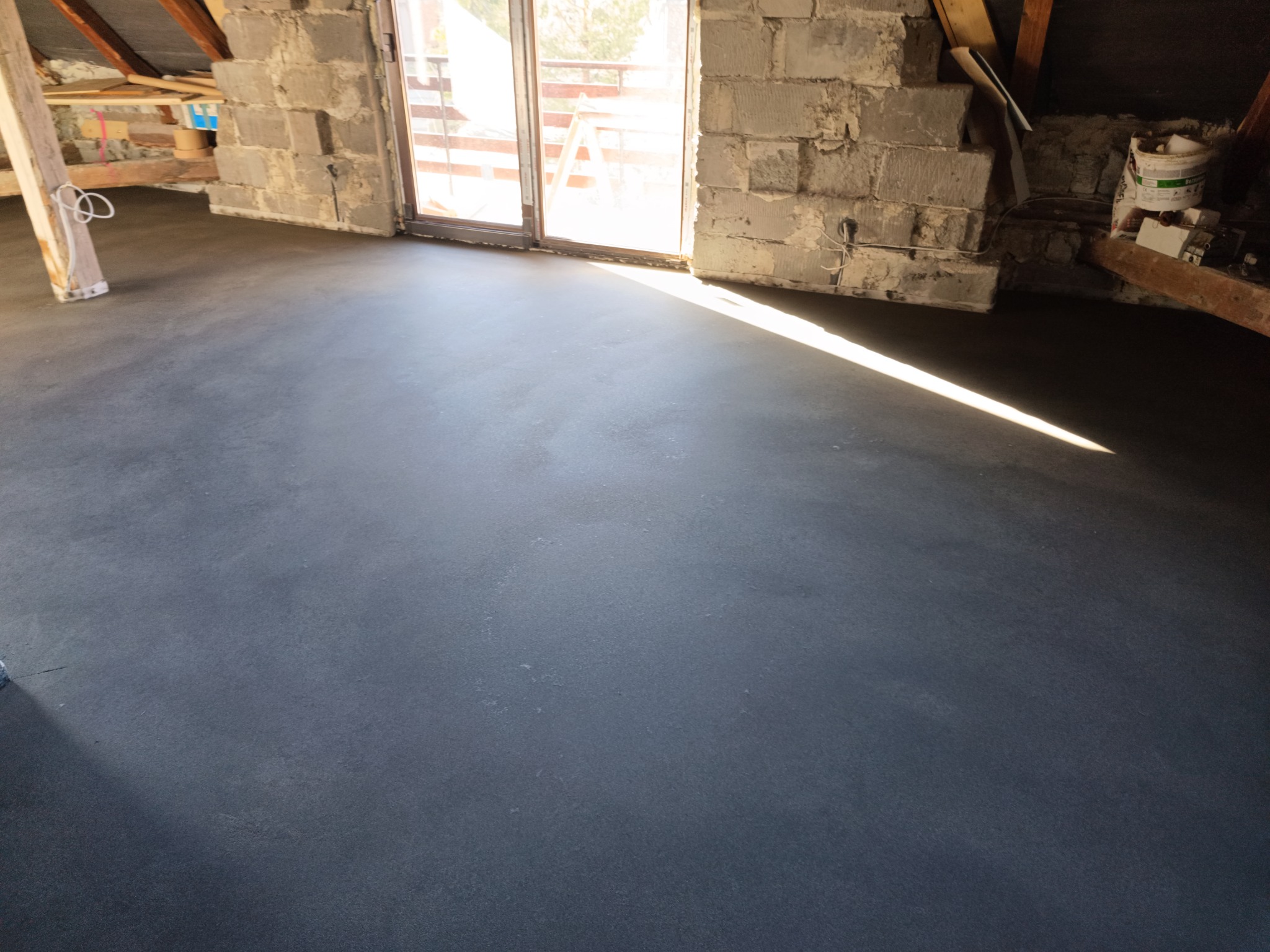 Świeżo wylana, szara posadzka w surowym pomieszczeniu na poddaszu, z widocznymi bloczkami betonowymi i fragmentem okna balkonowego wpadającego światła słonecznego.