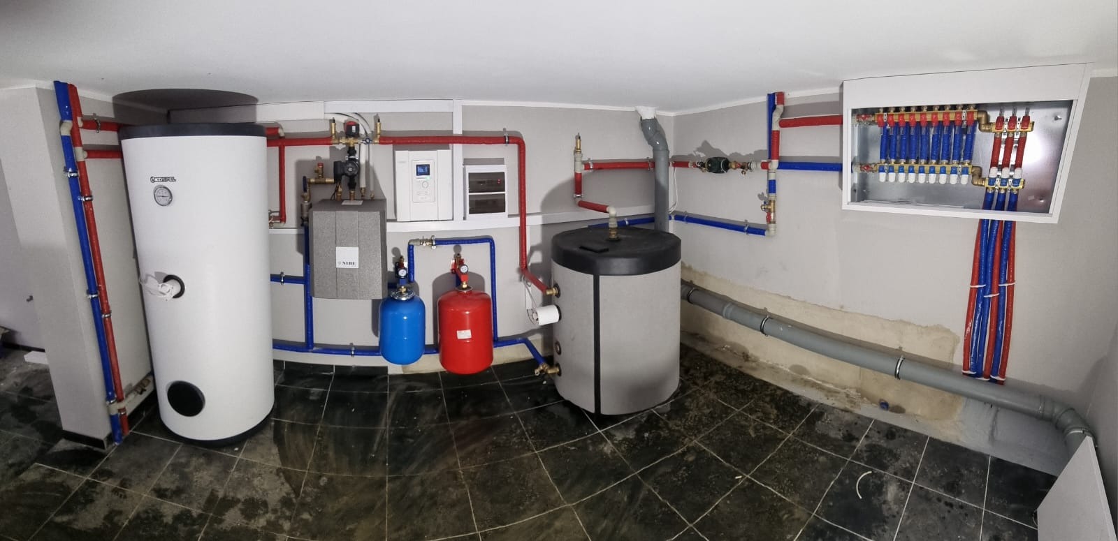 Kompletna kotłownia z dwoma zbiornikami, kotłem, naczyniami wzbiorczymi i rozdzielaczem podtynkowym, połączona rurami w kolorach czerwonym i niebieskim.