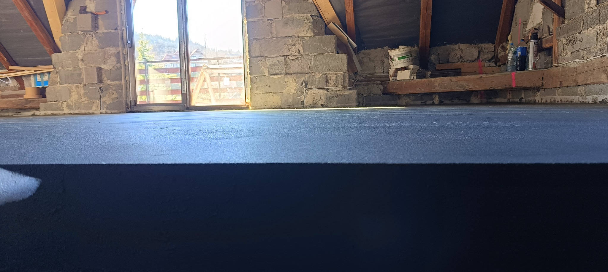 Gładka, ciemna wylewka na poddaszu w trakcie prac wykończeniowych, widok z niskiej perspektywy na tle okna balkonowego i surowych ścian z bloczków.