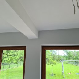 Nowi-Rem Paweł Nowicki - Wnętrze pokoju po remoncie: gładko pomalowany sufit z wystającymi belkami, dwa okna z drewnianymi ramami z widokiem na zielony ogród z huśtawką, wystające przewody elektryczne.
