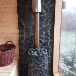 Nowi-Rem Paweł Nowicki - Wnętrze sauny z piecem opalanym drewnem obłożonym kamieniami, komin z metalu, ściana wyłożona kamieniem, drewniane ławki i koszyk wiklinowy.