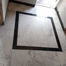 Ułożona podłoga z płytek imitujących marmur, obramowana czarnymi płytkami, widoczne pomarańczowe kliny dystansowe, surowy stan wykończenia ściany i widok na zewnątrz przez niedokończone drzwi.