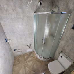 Nowi-Rem Paweł Nowicki - Narożna kabina prysznicowa z matowego szkła, toaleta i drewnopodobne płytki podłogowe w trakcie remontu łazienki; widoczne wystające kable elektryczne i rury doprowadzające wodę.